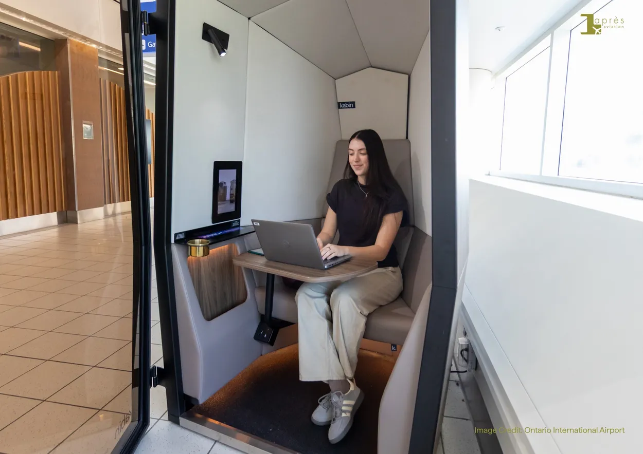 A traveller working on a laptop inside an open Kabin by Après Aviation pod beside a terminal window at Ontario International Airport.